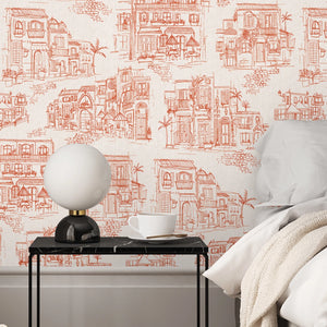 Charming Lanes wallpaper by Brand McKenzie in orange, toile-style townhouses behind a bed