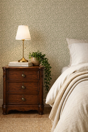 Elsie wallpaper by Curate Space in lichen green, dried poppy head motifs in a bedroom with a wooden bedside chest and table lamp